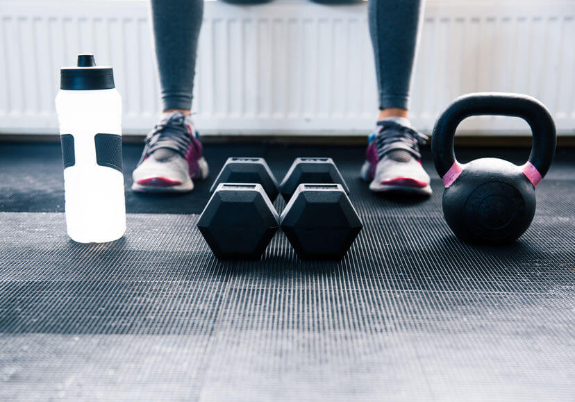 woman sitting at gym with dumbbells, protein shaker and kettlebell